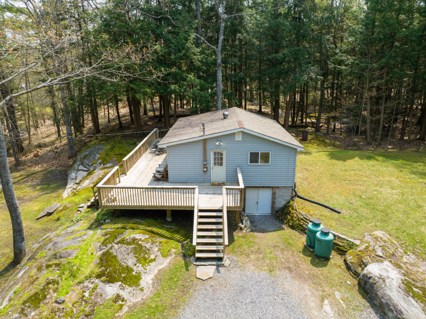 A small cottage with a large wrap-around deck sits in the centre of a clearing surrounded by trees.