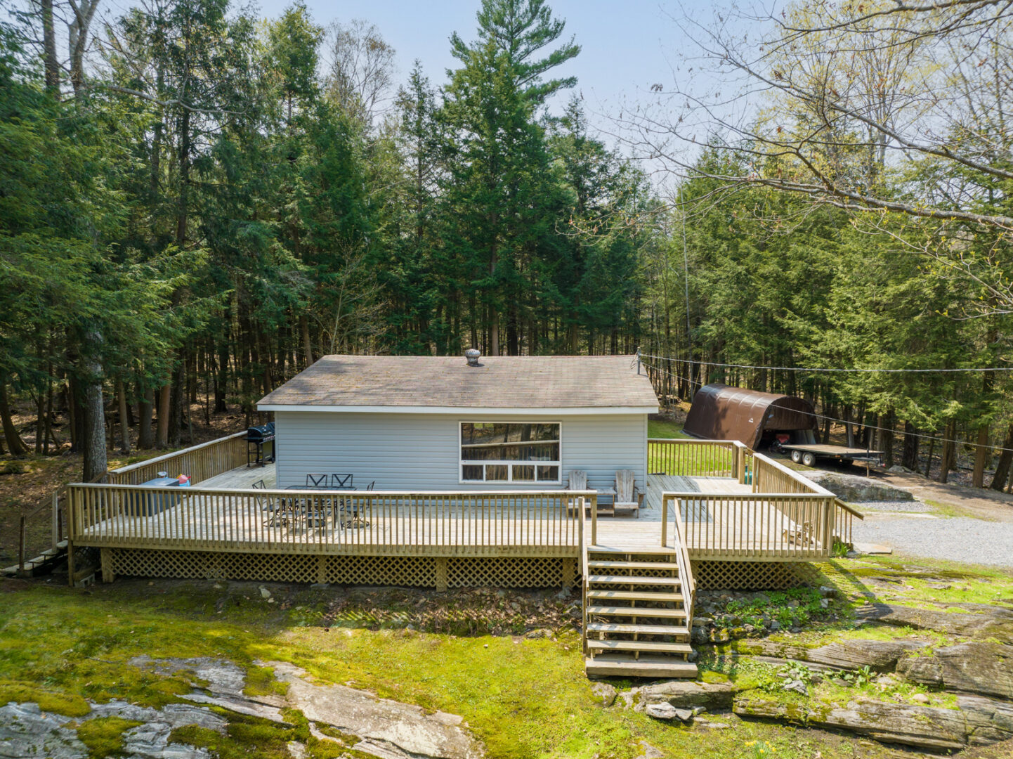 A small cottage with a large wrap-around deck sits in the centre of a clearing surrounded by trees.