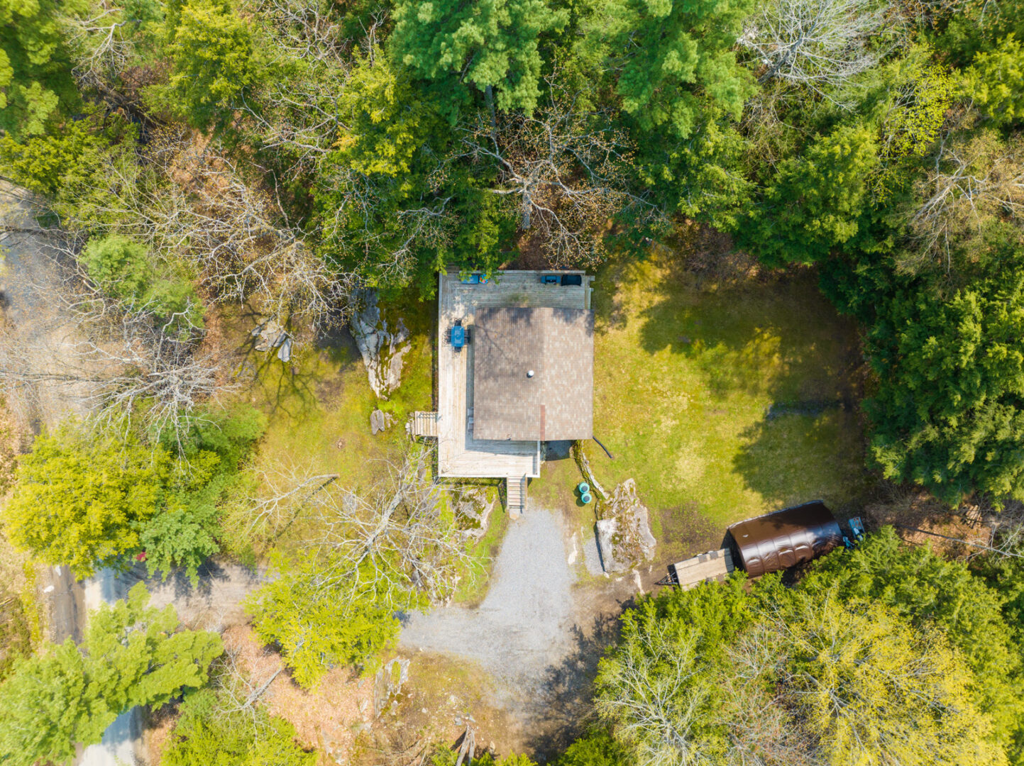 Aerial view of a small cottage with a large wrap-around deck that sits in the centre of a clearing surrounded by trees.