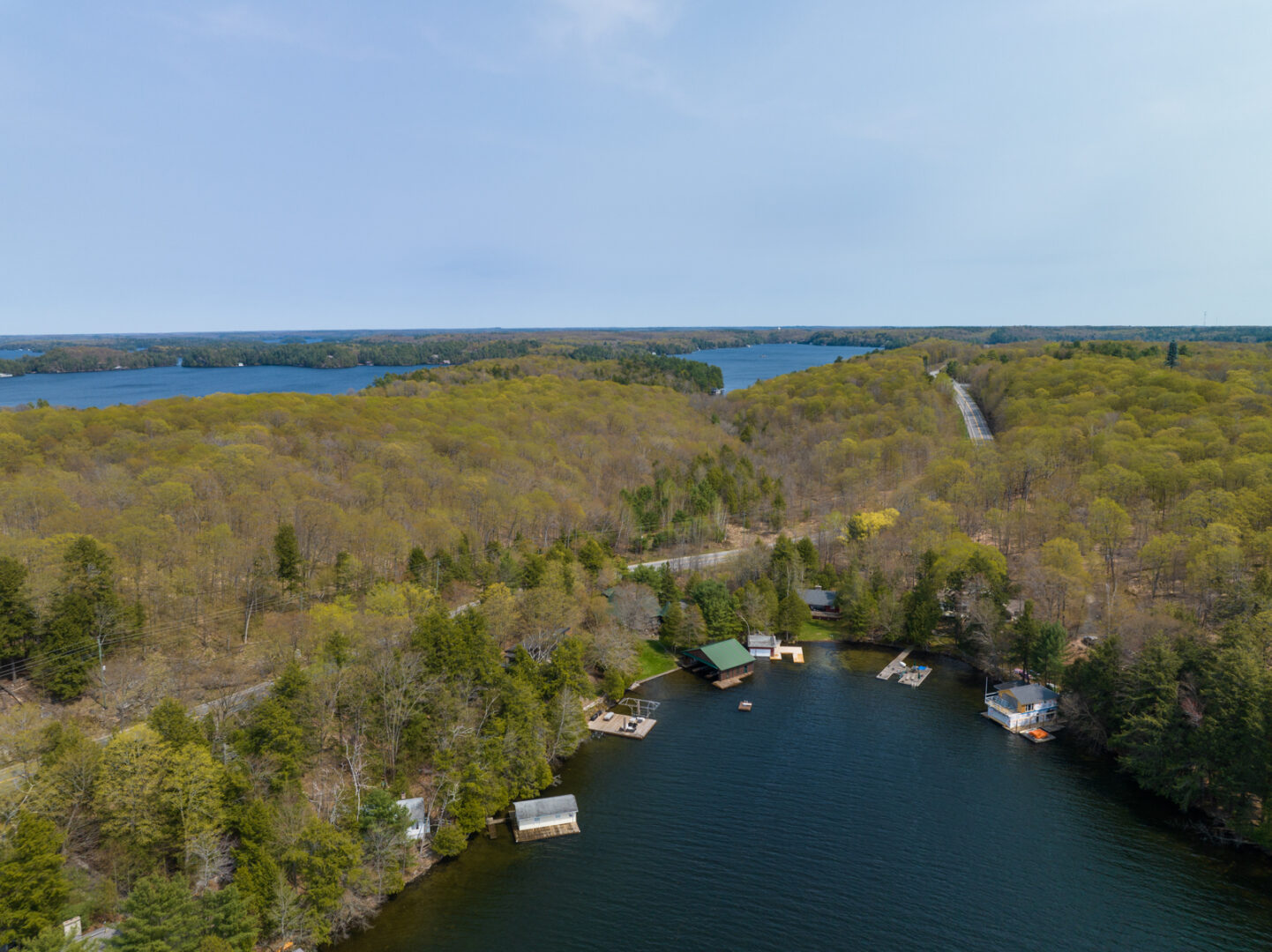 Cottages line a curved shoreline of a lake. Thick trees stretch out behind and more lake can be seen beyond.