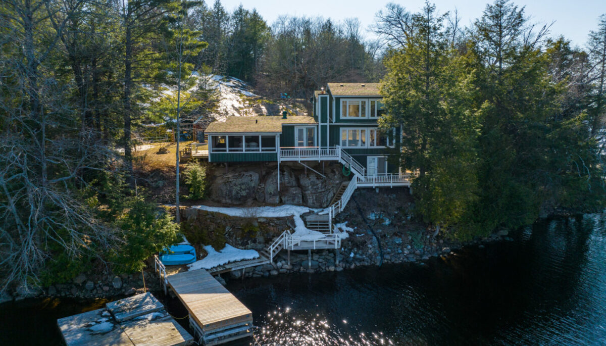 A large cottage with lots of windows sits on the shoreline of a lake. A large dock extends into the water.