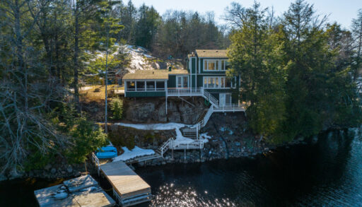 A large cottage with lots of windows sits on the shoreline of a lake. A large dock extends into the water.