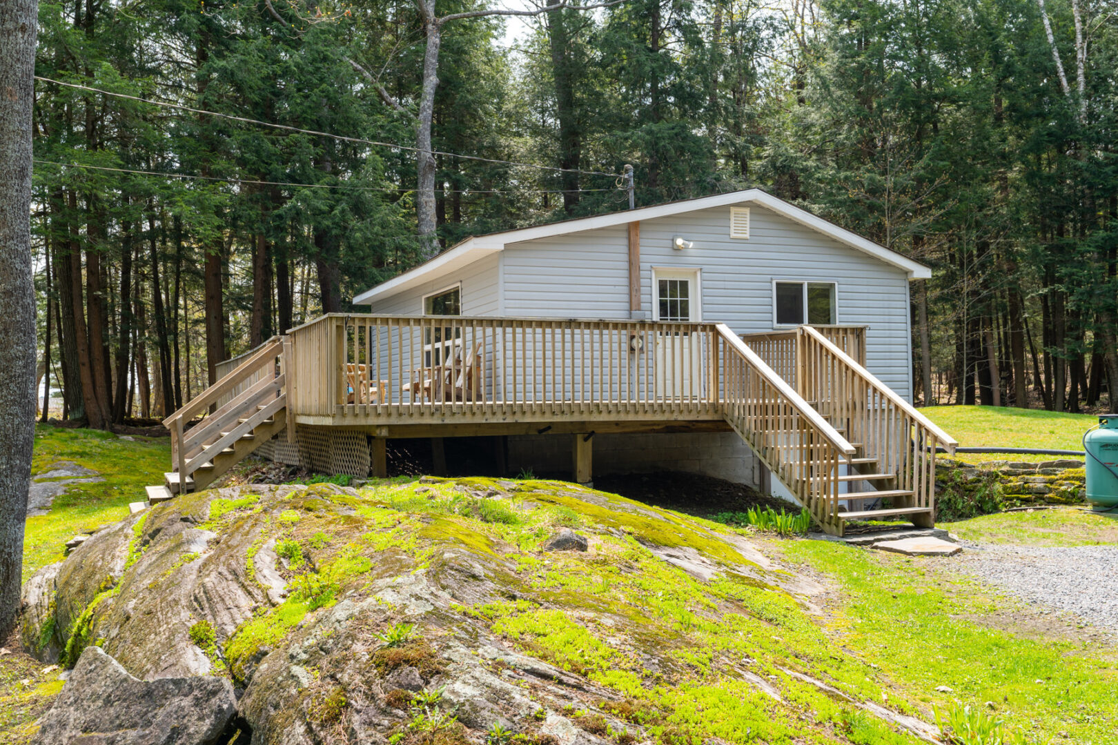 Exterior of a small cottage with a large deck. The sloping front yard is a mix of large rocky ground and grass.