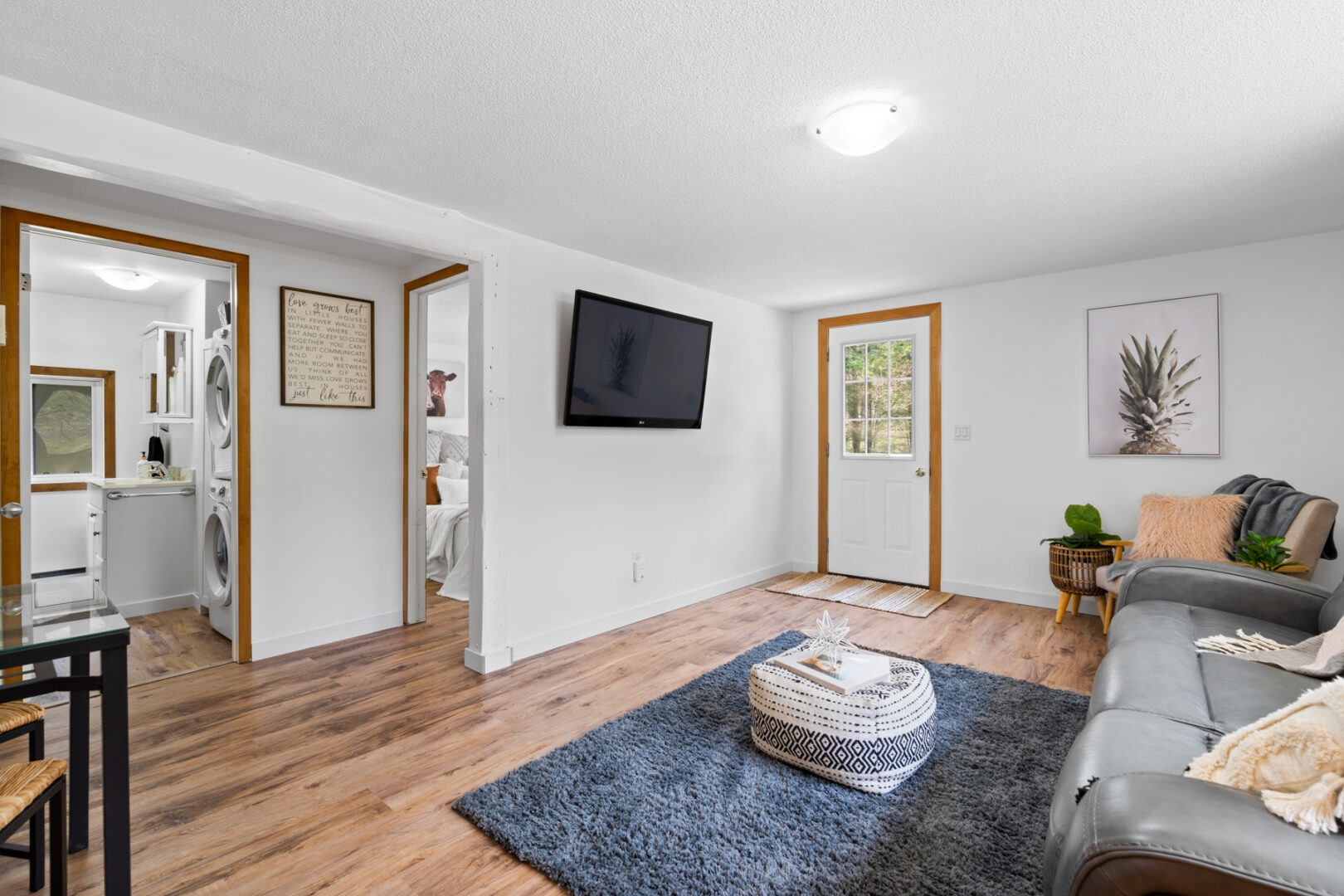 Inside of the front door of a small cottage, leading straight into a bright living area with a rug, a couch, and a television. Doorways off to the side lead to a washroom and a bedroom..
