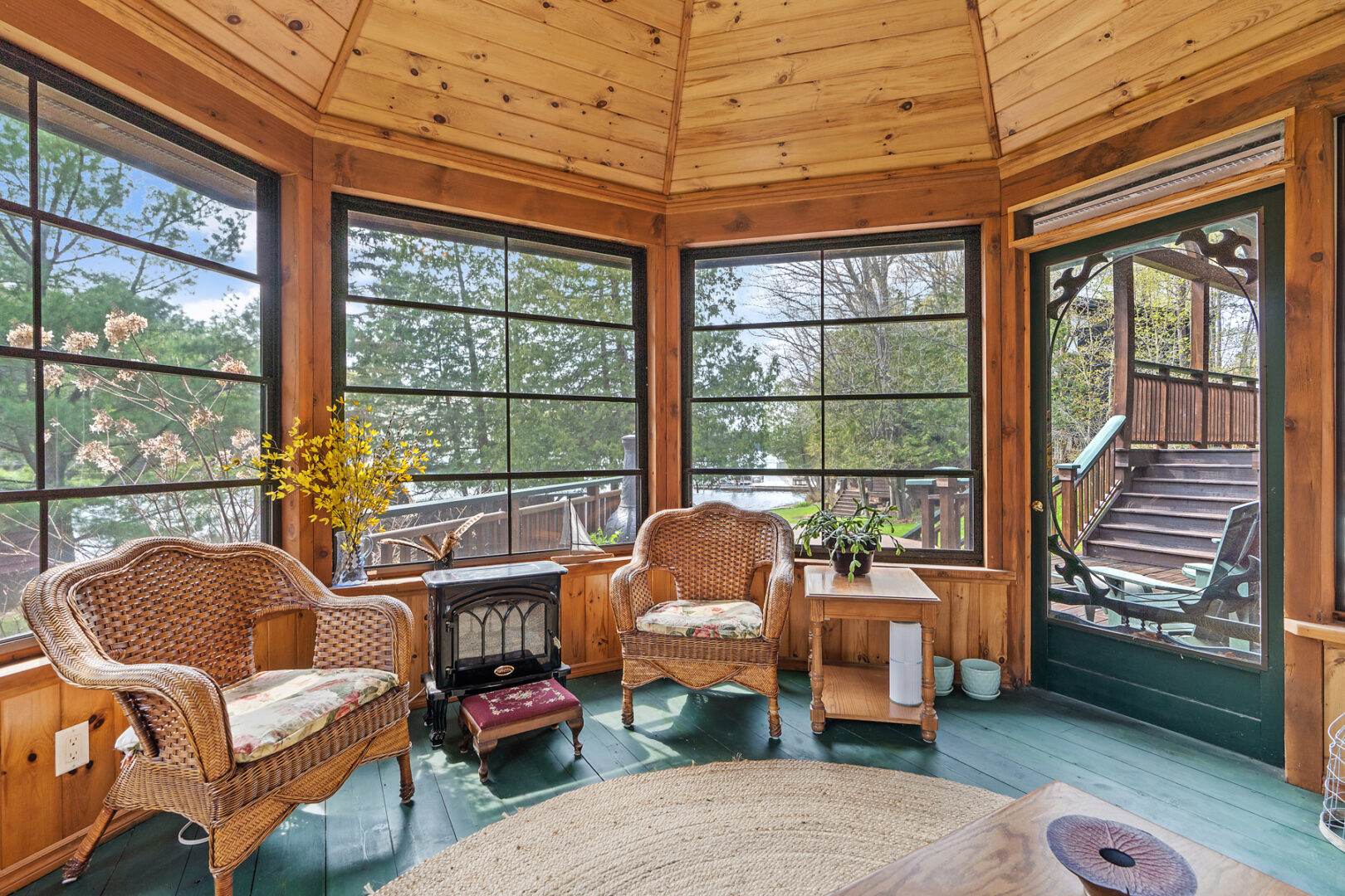 Interior of an octagonal screened-in room with tons of seating, a wood-panelled ceiling, a ceiling fan, and a screen door leading to a back deck.