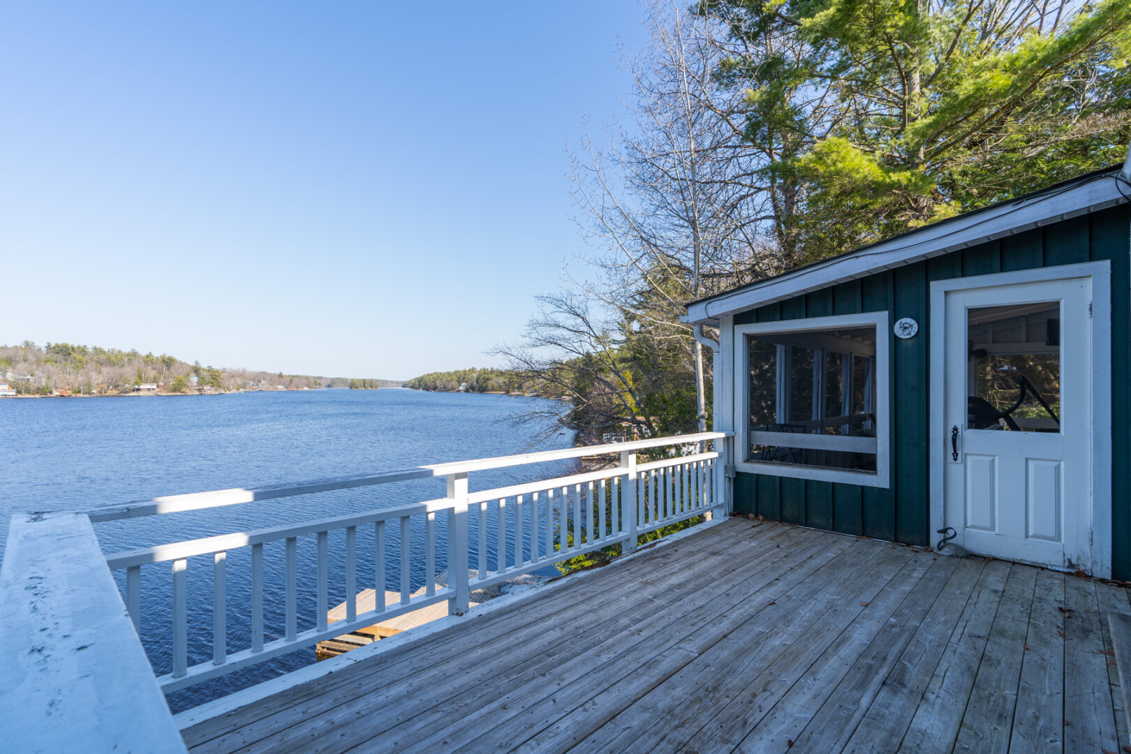 A large deck with a white railing extends off the side of a cottage, overlooking a lake.