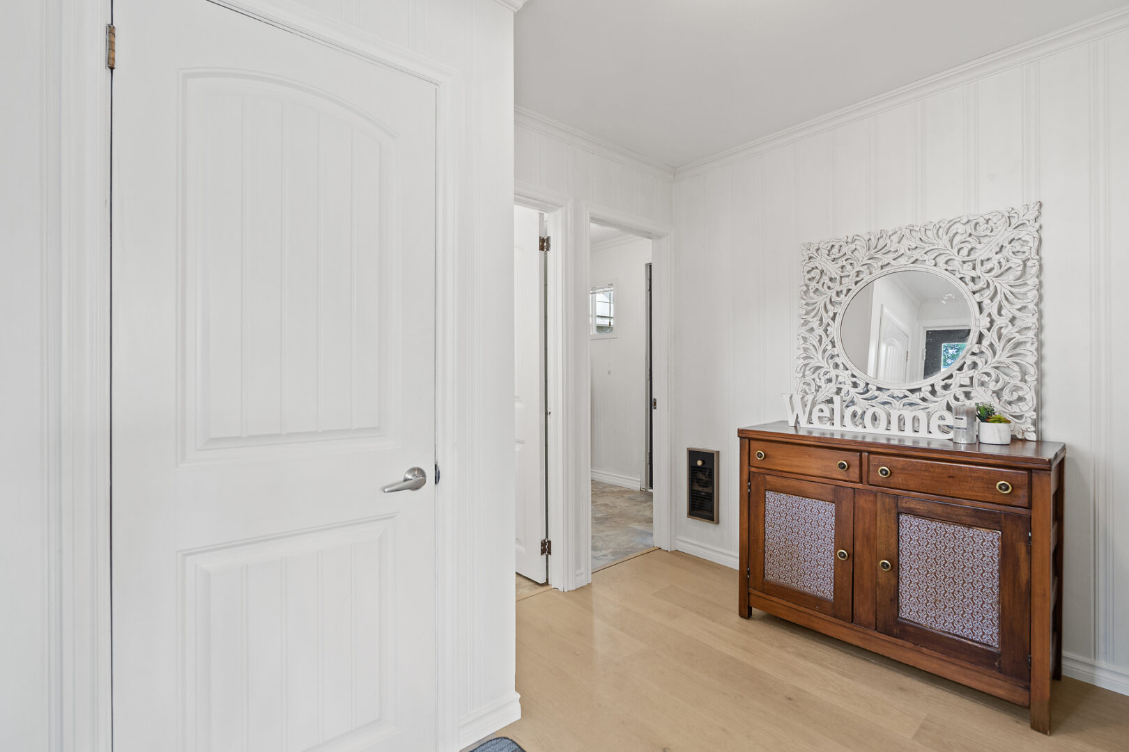 A bright but narrow entrance hall of a cottage, with shite walls, hardwood floors, and a "welcome" sign.