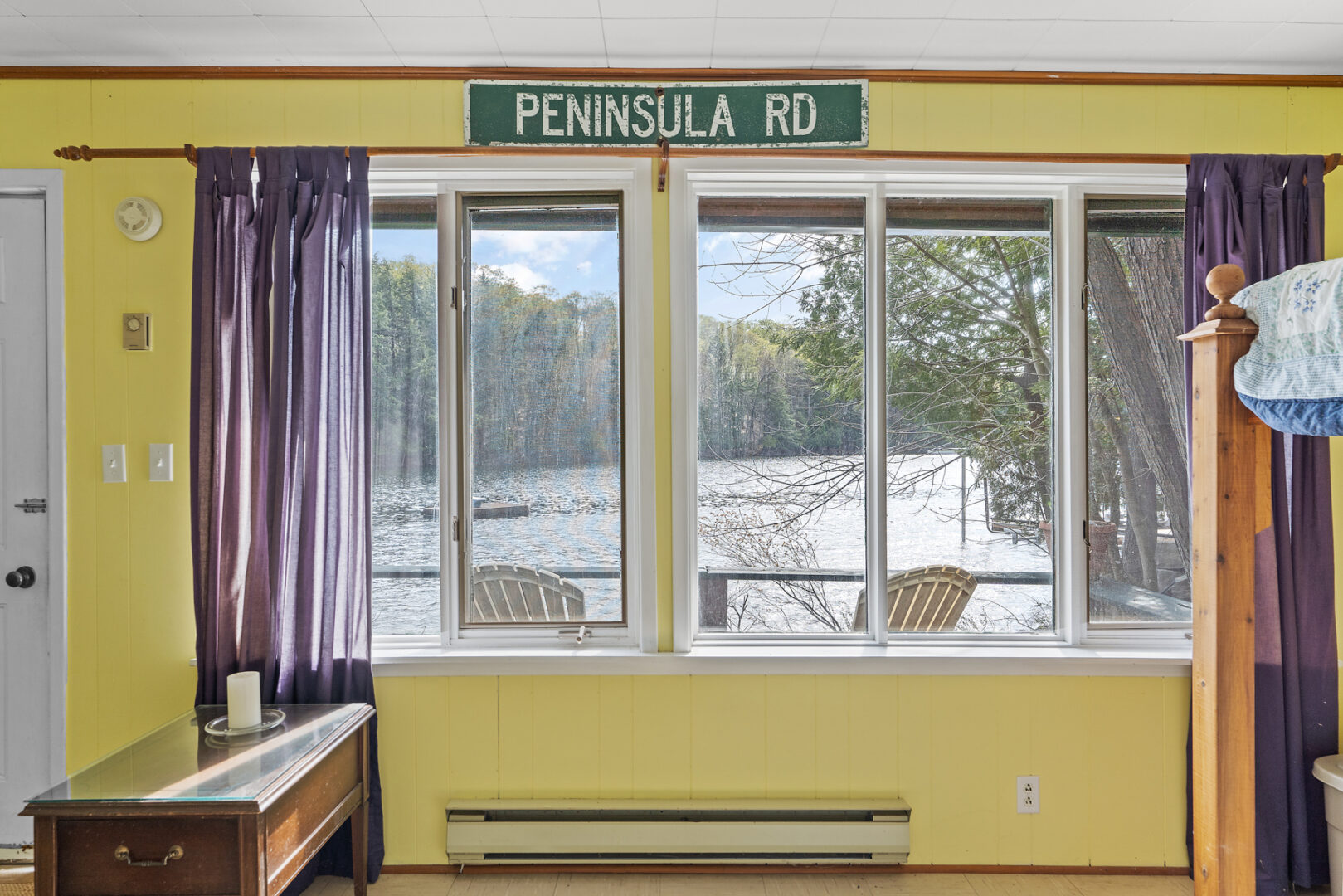 The view from big windows inside a small bunkie, looking out to the lake. A sign above the window reads "Peninsula Rd."
