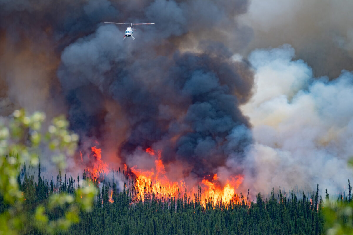 Fire spreads across a Canadian forest in the distance, smoke billows upward as helicopter flies away