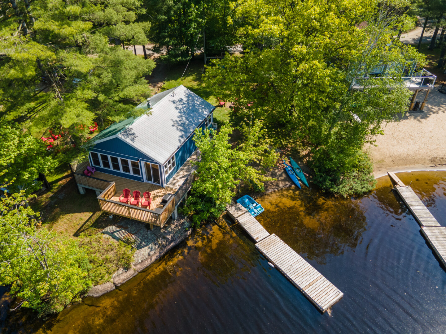 Overhead view of a small blue cottage with a big deck, a sandy shore, and a long dock.