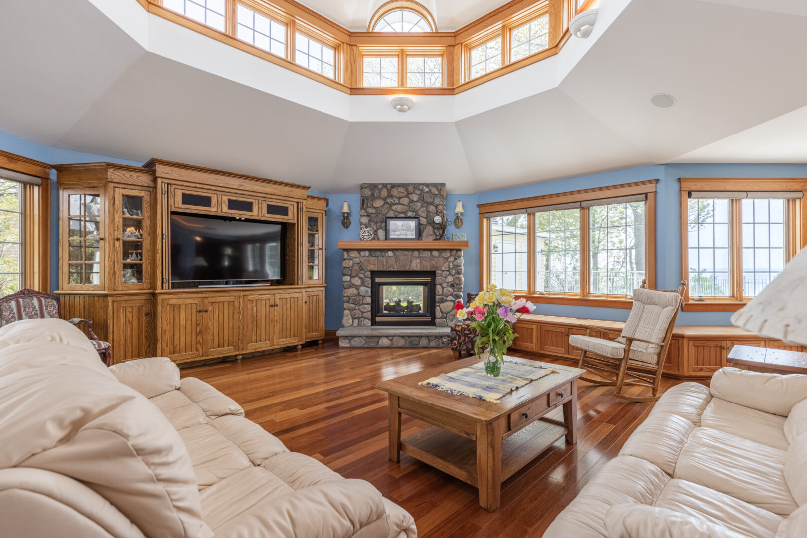 A large lake house living area with a big sectional couch, hardwood floors, a television, and a stone fireplace.