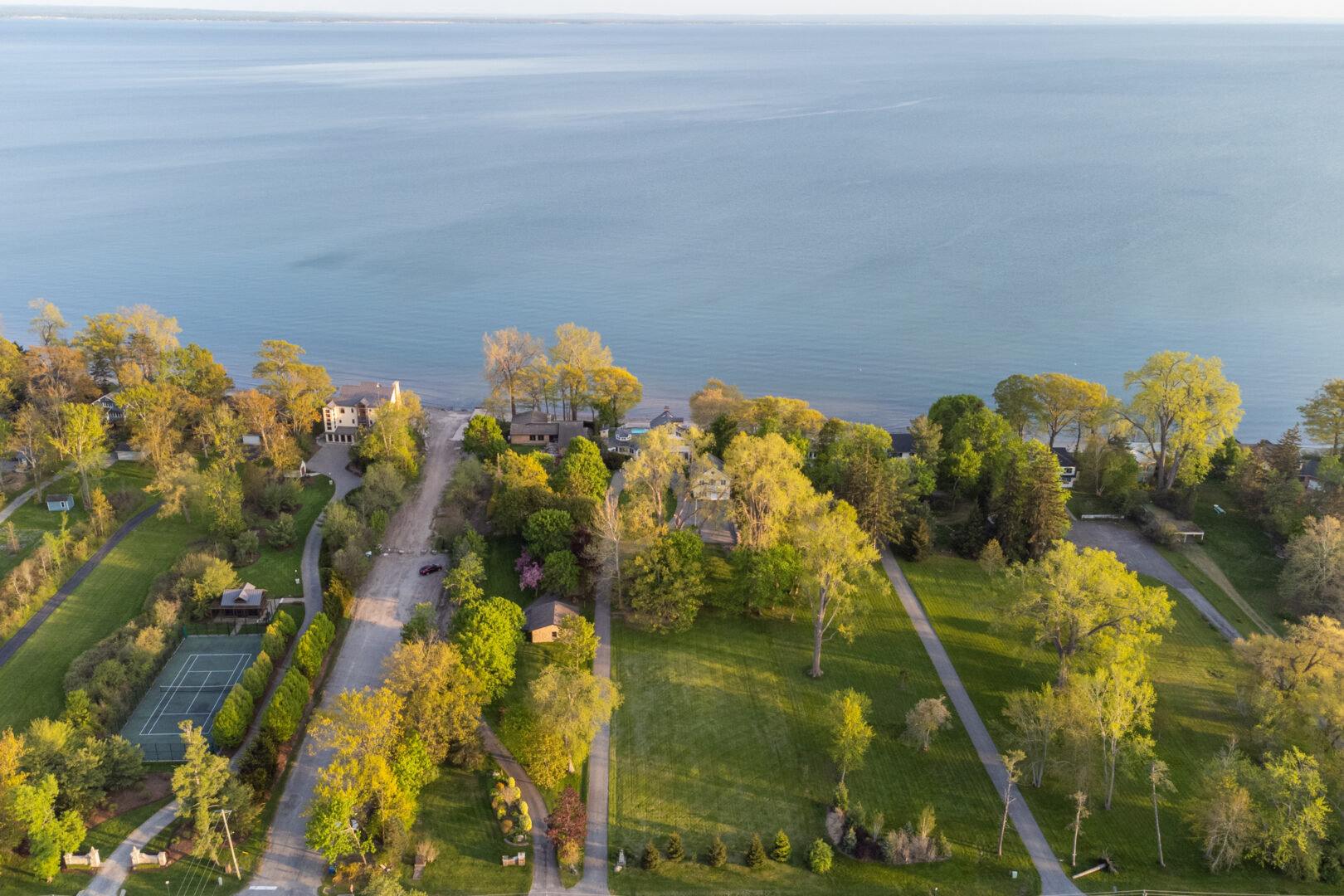 Overhead view of a large area of land in front of a lake. A long, narrow green lawn leads to a big lake house surrounded by trees.