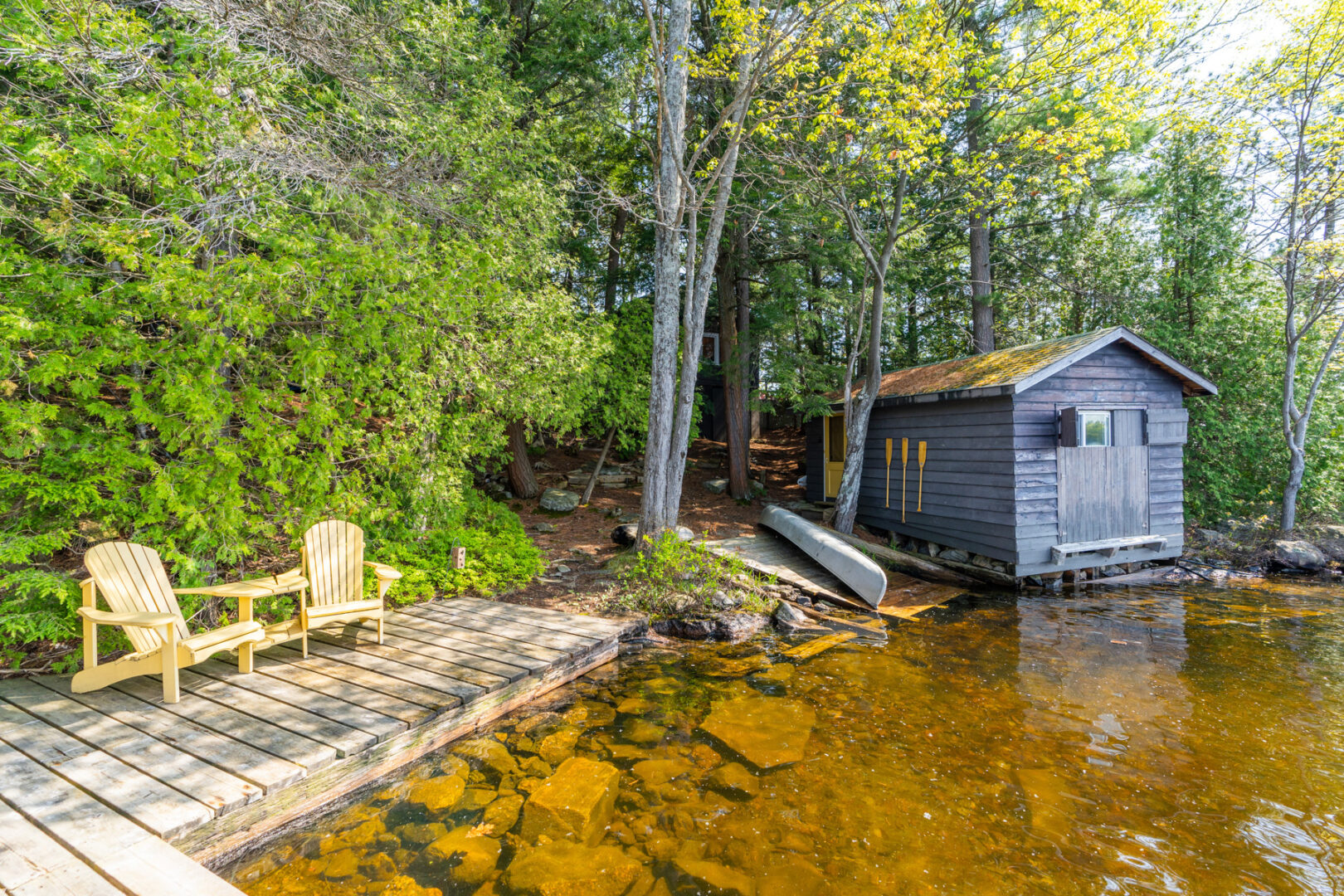 A stretch of lake shoreline with a small dock and two yellow Muskoka chairs, and a bunkie sitting along the water's edge.