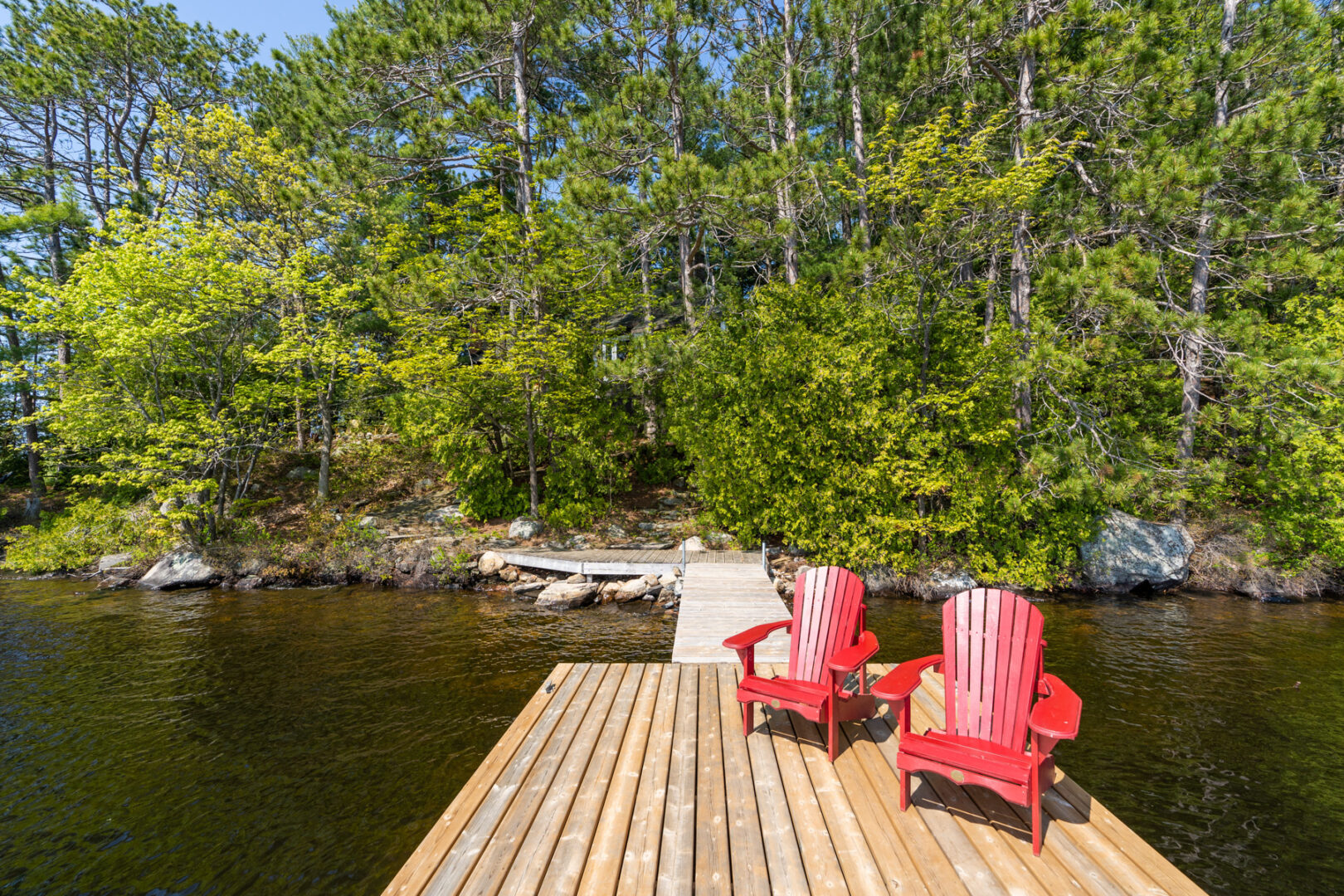 A long dock with two red Muskoka chairs extends into a blue lake from a shoreline of green trees.