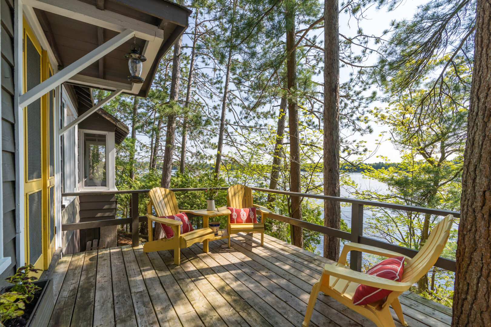 A large deck extending off the back of a cottage, with a set of yellow Muskoka chairs and a lake view.