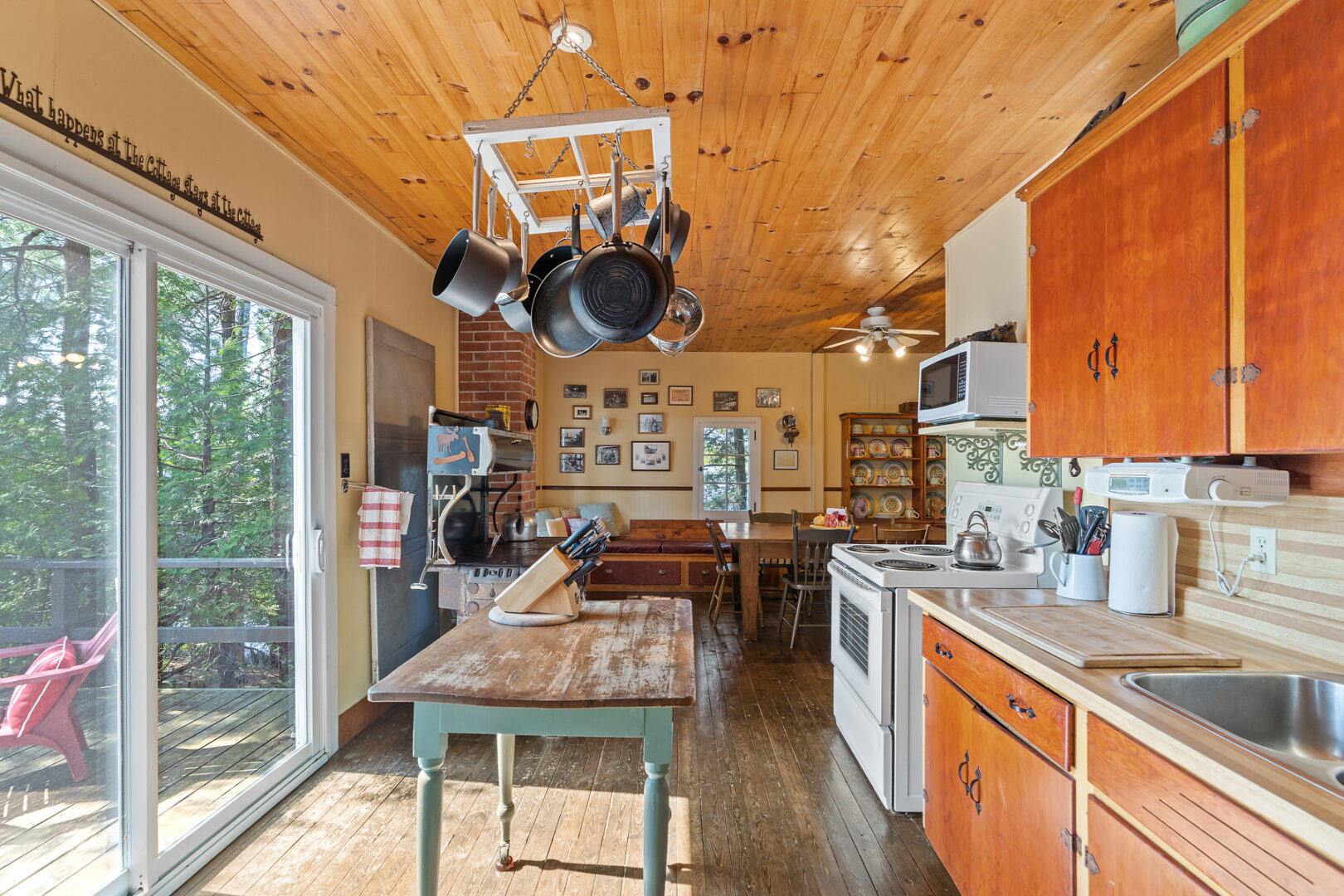 A small cottage kitchen with wooden cabinets, an oven, pots and pans hanging from the ceiling, double doors opening to a deck, and a table..