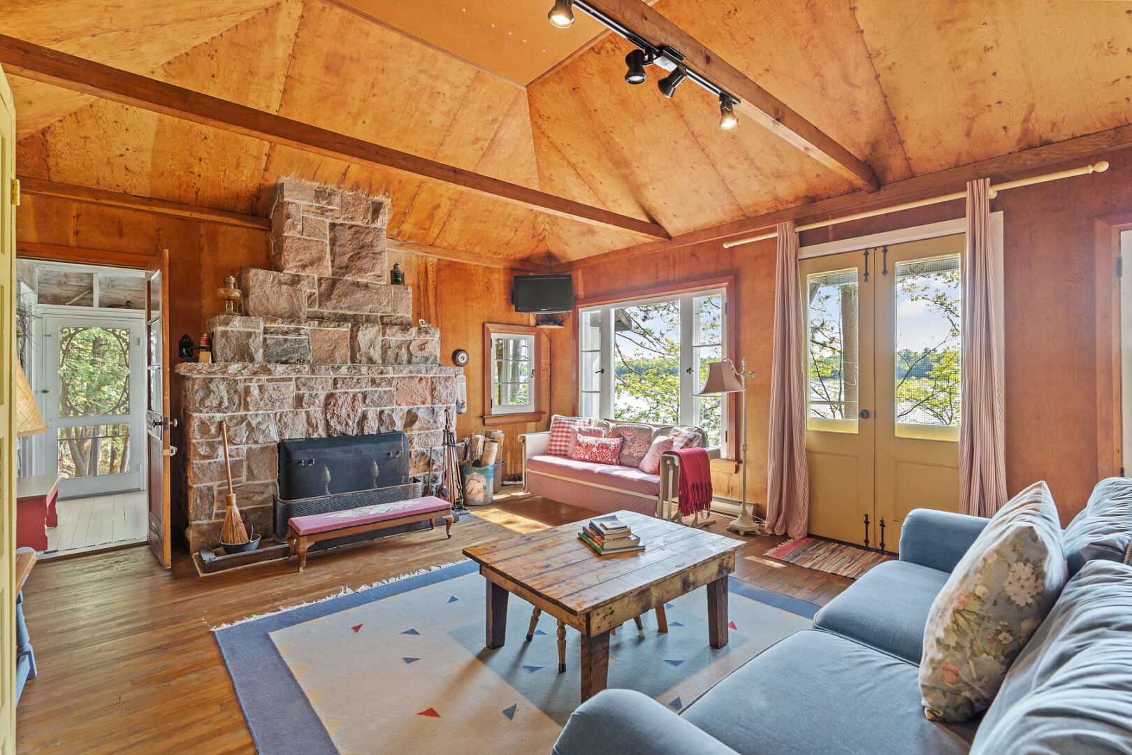 A bright, simple cottage living area with large windows, a couch, and a stone fireplace.