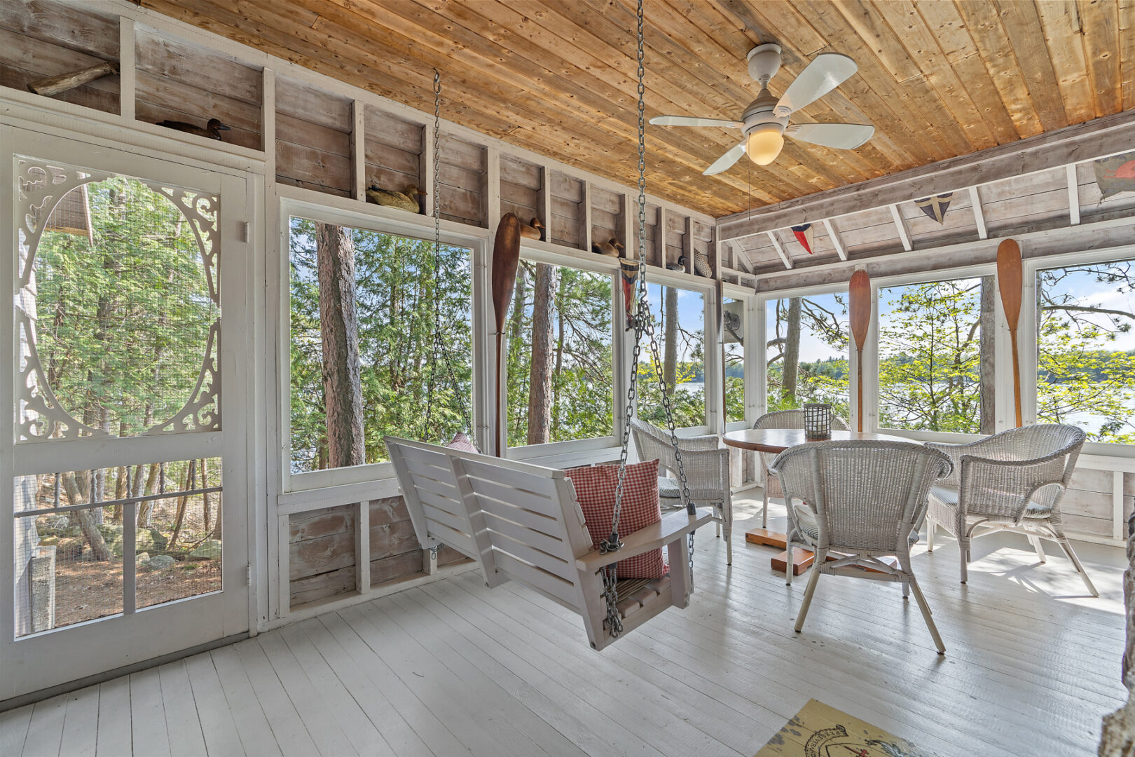 A bright cottage sunroom with white deck flooring, a white swing, and white wicker furniture.