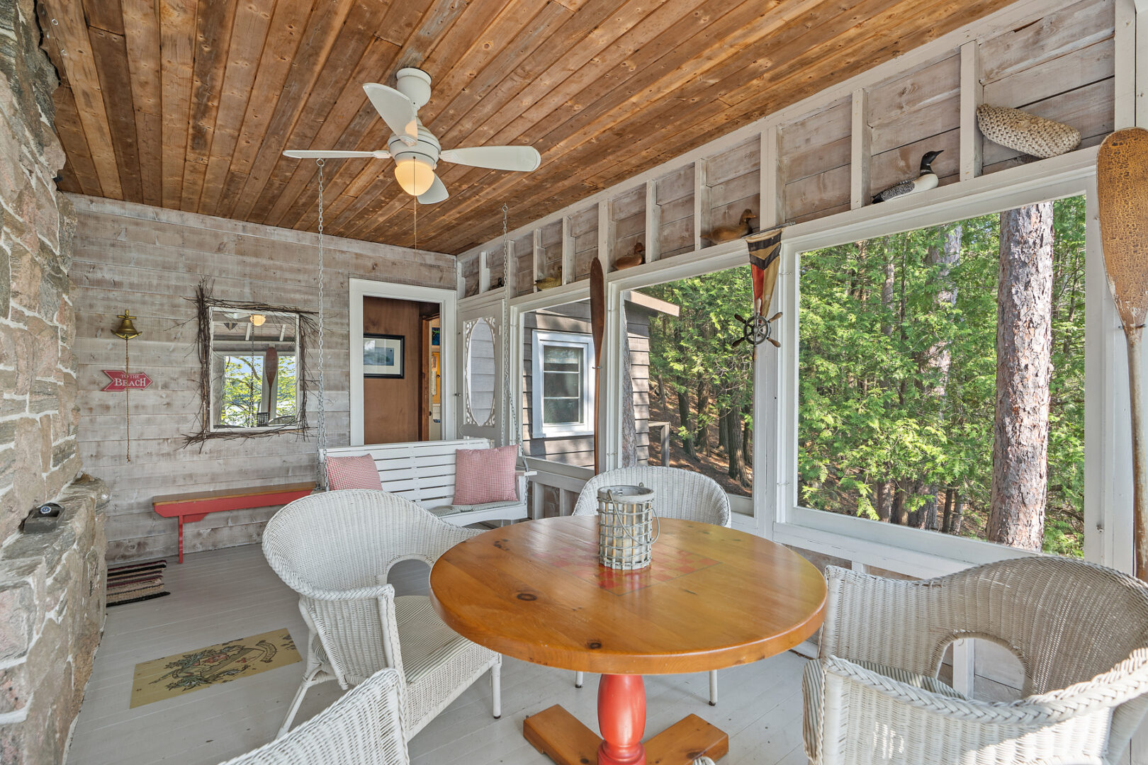 A bright cottage sunroom with white deck flooring, a white swing, and white wicker furniture.