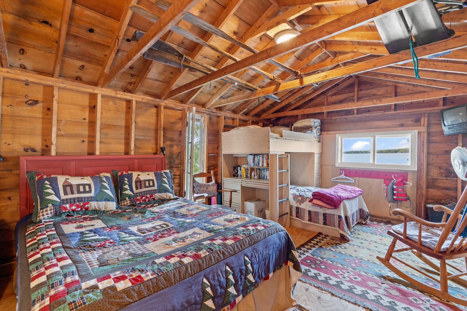 Interior of a lakeside bunkie with wooden beams stretching across a slanted ceiling, a large bed, a set of bunkbeds, and a small window.