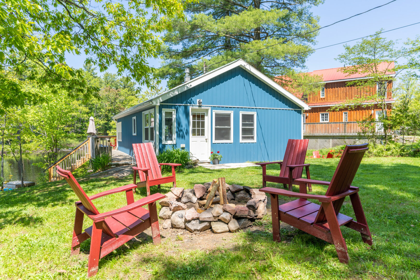A fire pit sits in the middle of a green yard, surrounded by four red Muskoka chairs. A small blue cottage sits in the background.