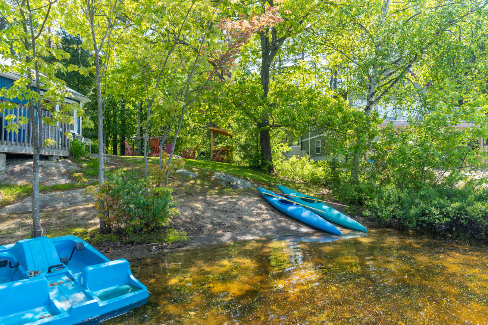 A small, shady, sandy stretch of shoreline on a calm lake. Two blue kayaks and a blue paddle boat sit on the shore.