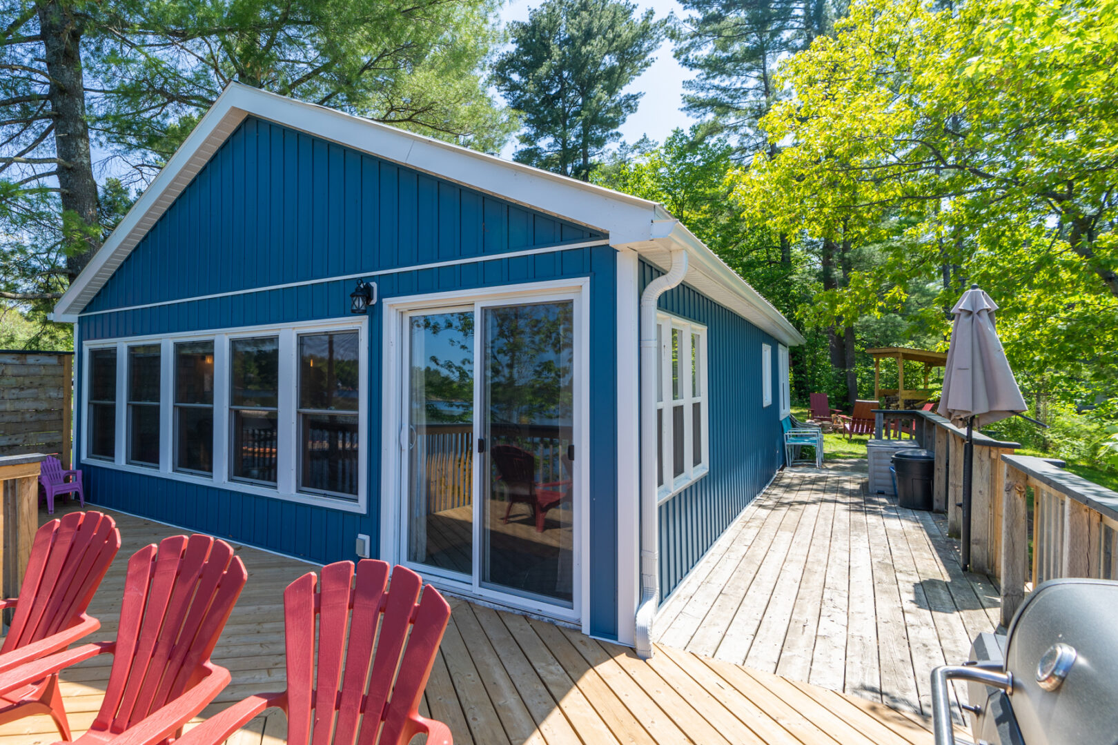 Lake-facing side of a small blue cottage with lots of windows. A large deck with red Muskoka chairs wraps around two sides of the cottage.
