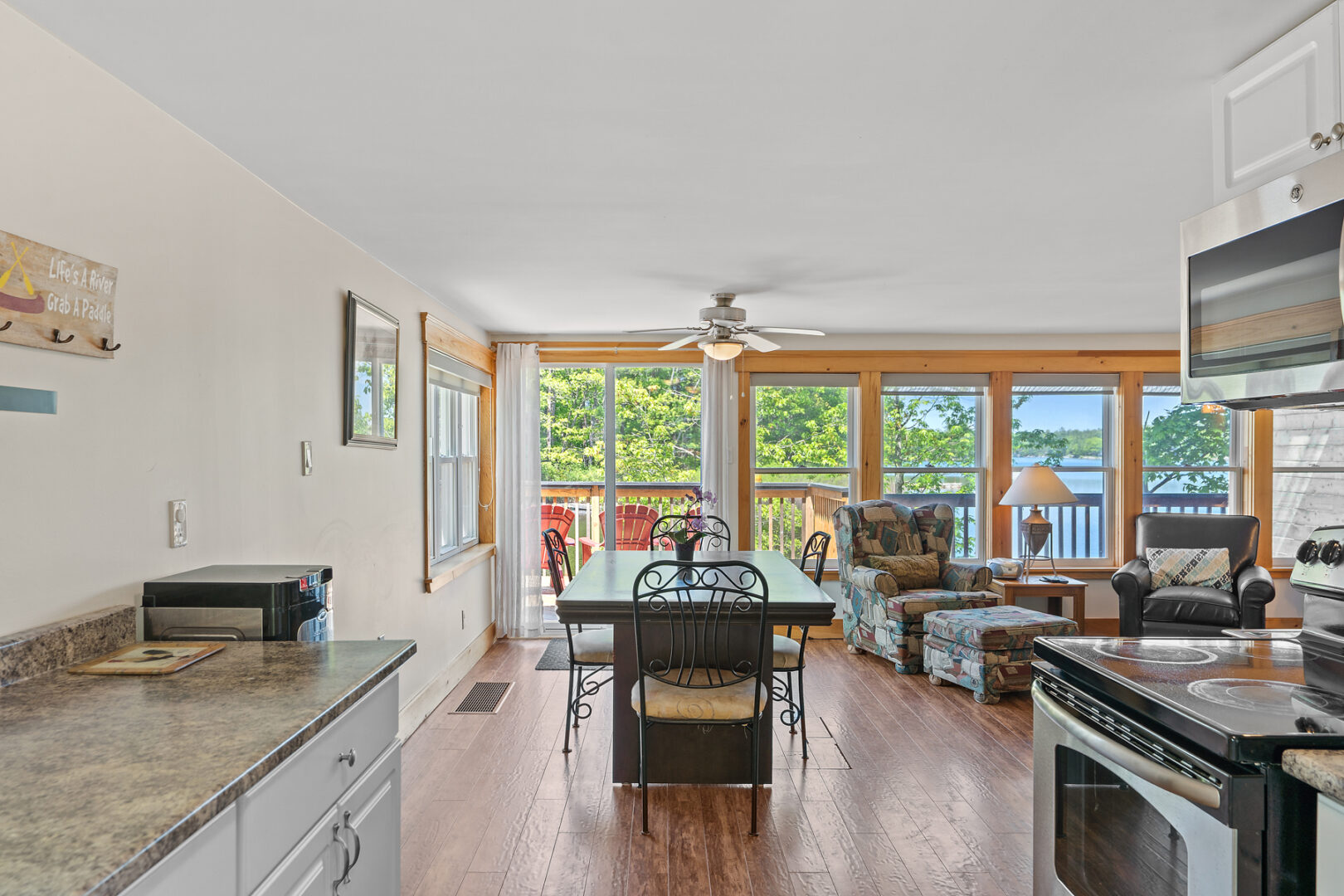 Inside the main living area of a small cottage. An open-concept kitchen flows into a living and dining area, with big windows looking out to a back deck.