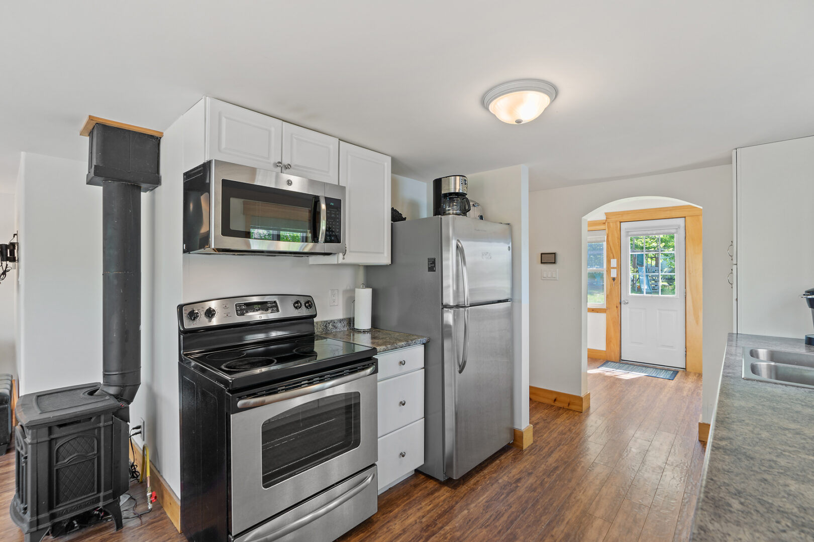 A small, bright kitchen space in a small cottage. An updated oven, microwave, and fridge sit nestled between white cabinets.