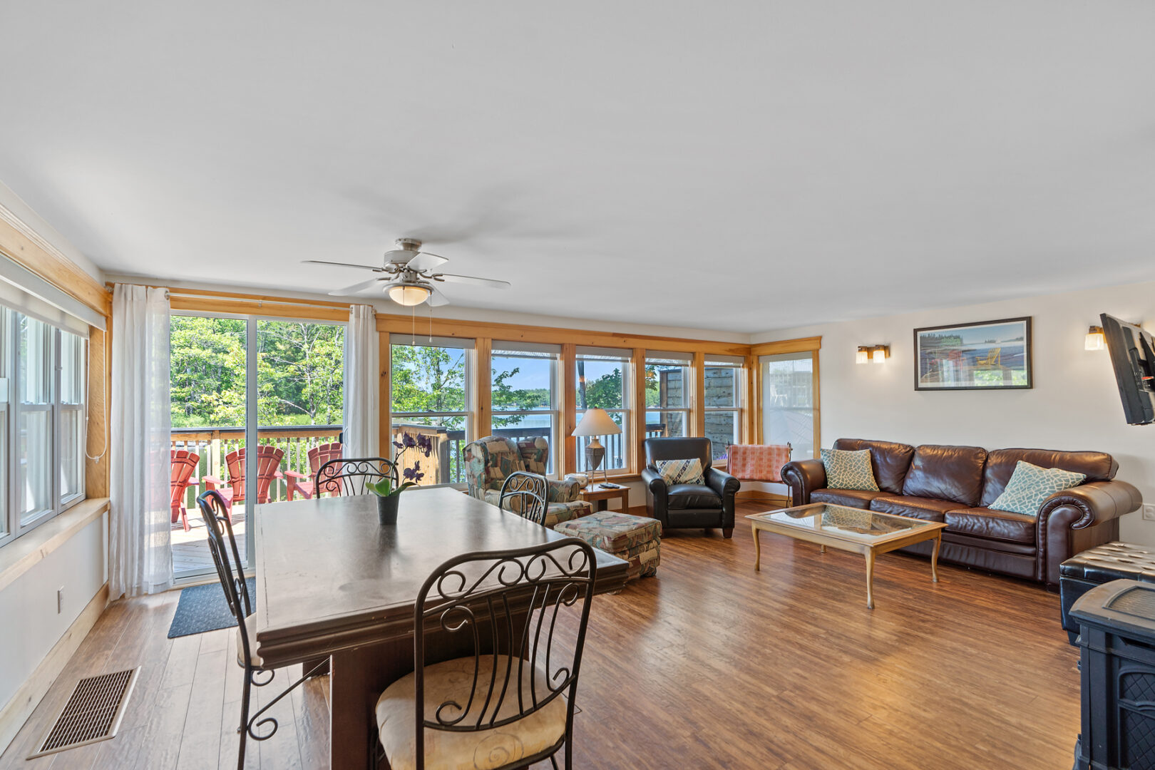 A bright, open-concept cottage living space with a dining table, couches, and big windows looking out to a back deck.