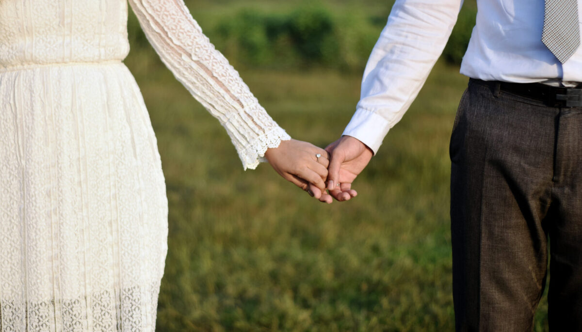 Formal attire romantic couple holding hands together while wearing engagement or wedding rings with their hands in a beautiful nature soft blur background illuminated by the gorgeous sun.
