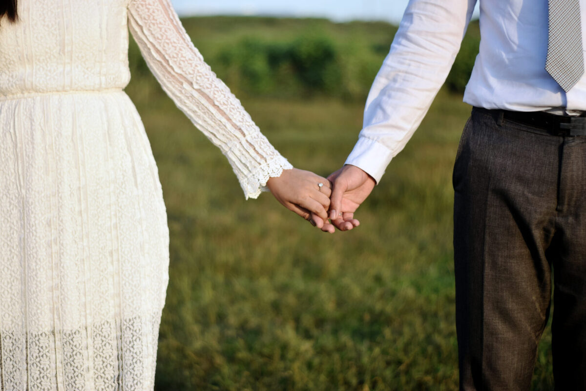Formal attire romantic couple holding hands together while wearing engagement or wedding rings with their hands in a beautiful nature soft blur background illuminated by the gorgeous sun.
