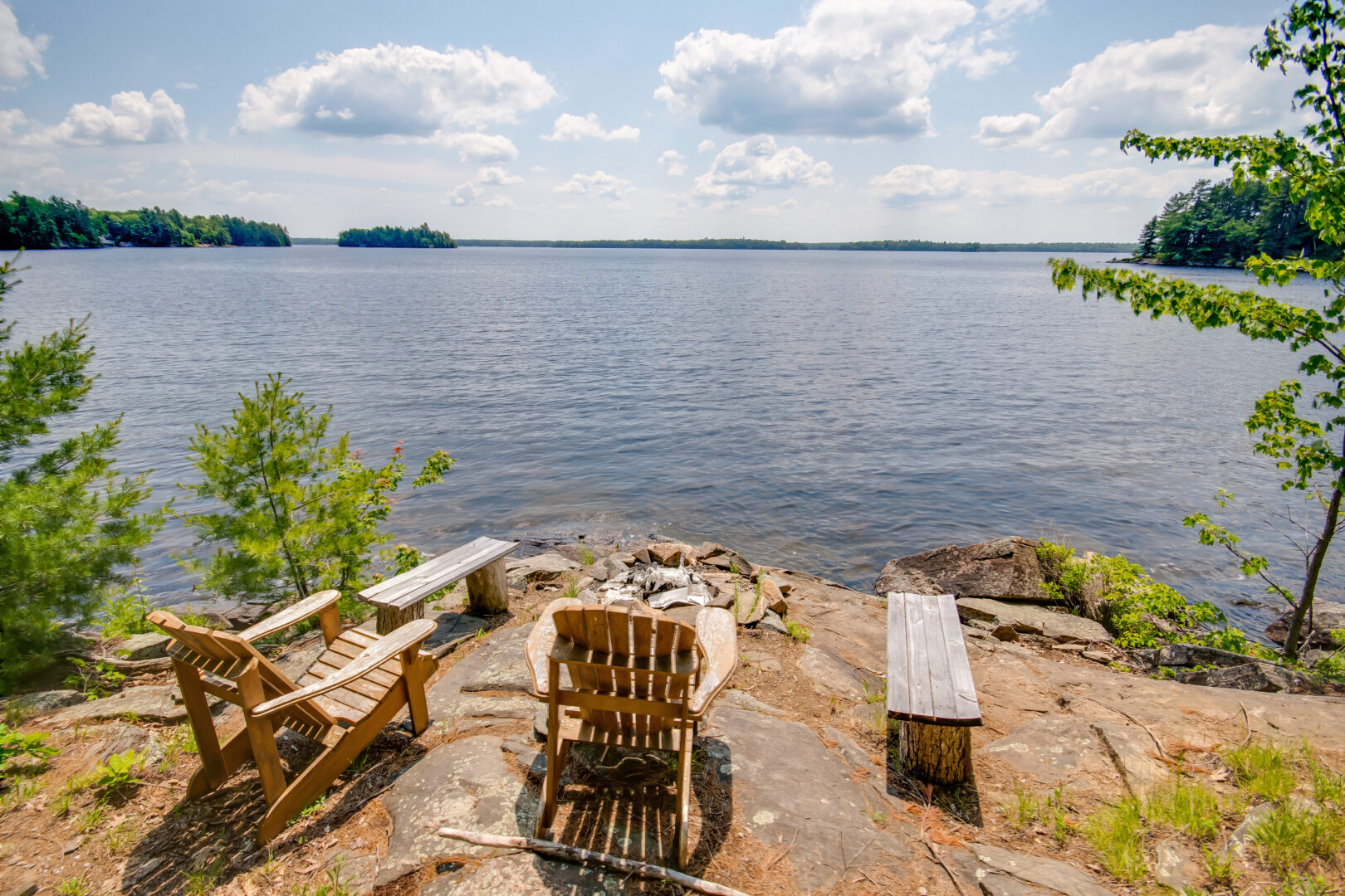 Two wooden chairs and two benches sit around a fire pit, overlooking a lake.