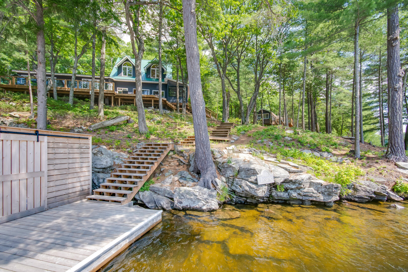 A cottage sits at the top of a sloping hill. A shallow waterfront with a dock sits at the bottom of the treed property.