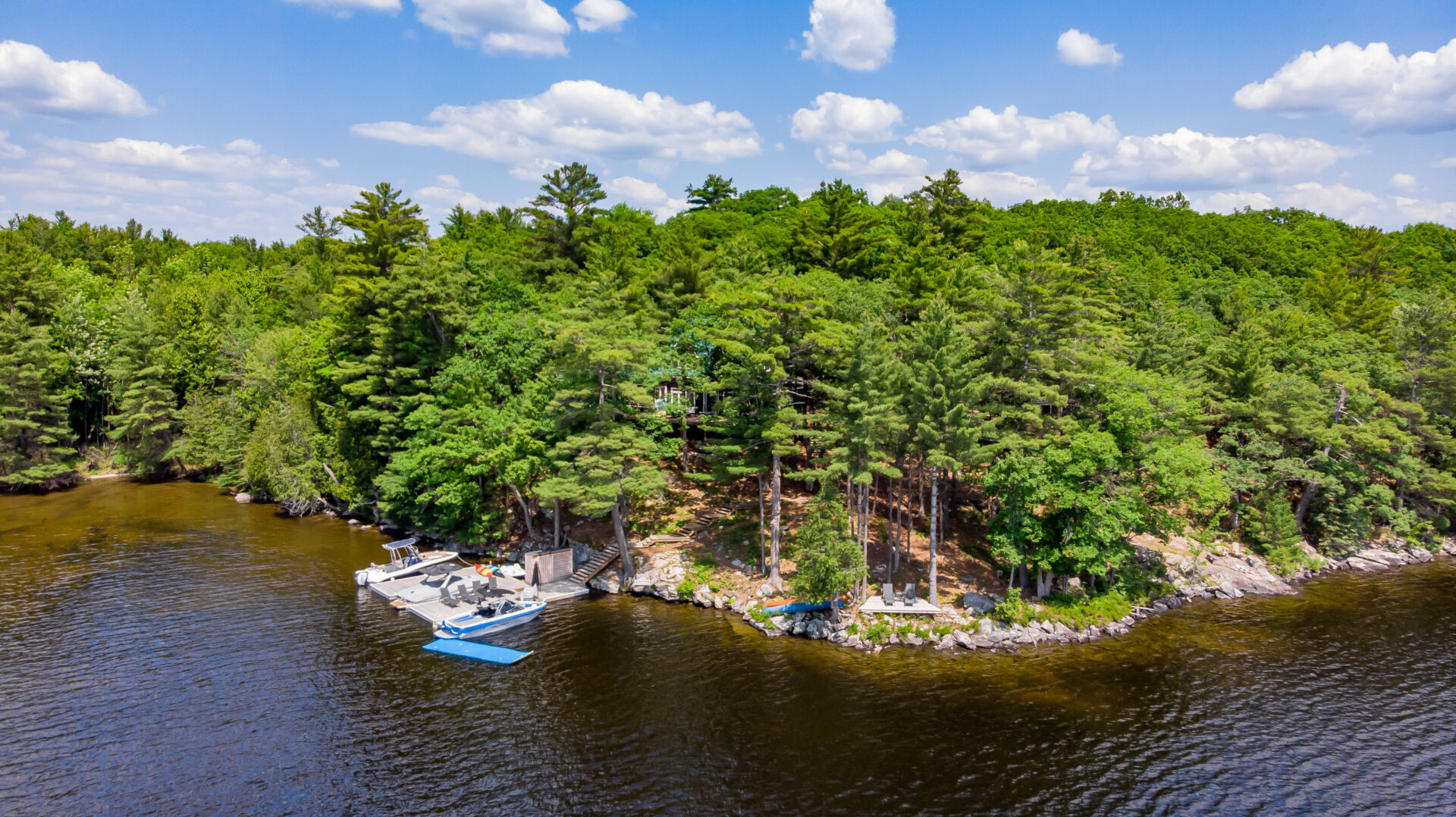 A stretch of shoreline curving out into a lake. A dock with a couple boats and another, separate dock with chairs looking out over the water line the shore.
