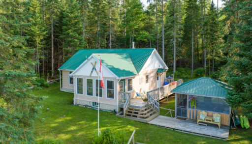 A cottage with big windows and a green roof sits on a grassy property. Beside it, there is a large, detached screened-in gazebo, also with a green roof.