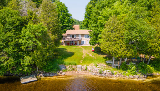 A house sits up the hill from the sandy shoreline of a lake, surrounded by trees.