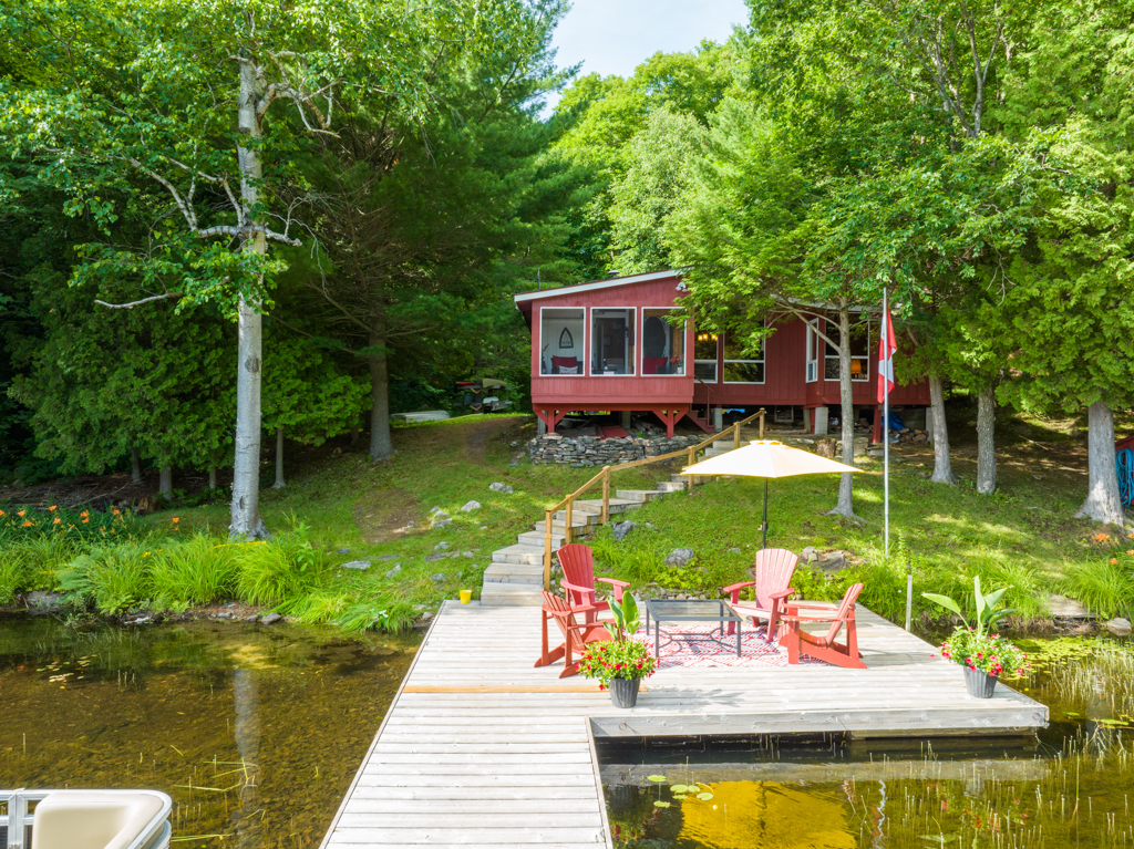 A small red cottage sits just up the shore of a lake, surrounded by green trees. A dock stretches out into the lake in front.