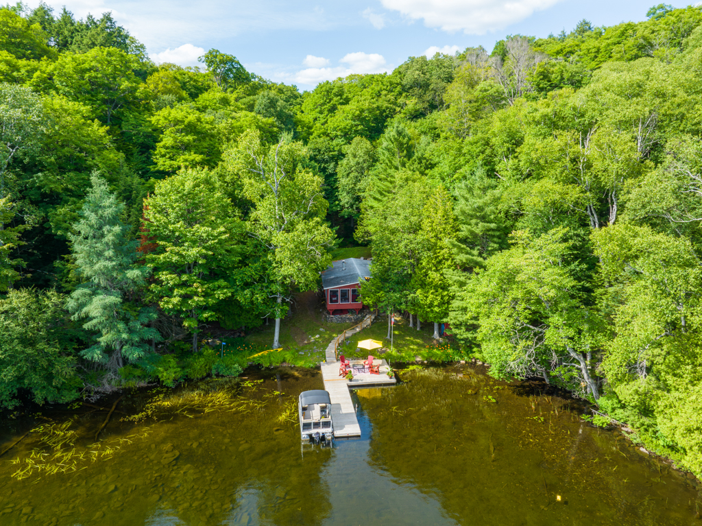 A cottage sits tucked into the shoreline of a small, curving lake bay, surrounded by green trees.