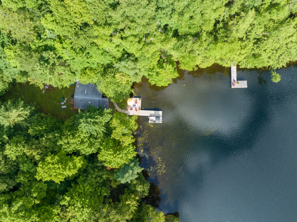 Overhead view of a cottage surrounded by green trees, with a dock that stretches into the bay of a lake.