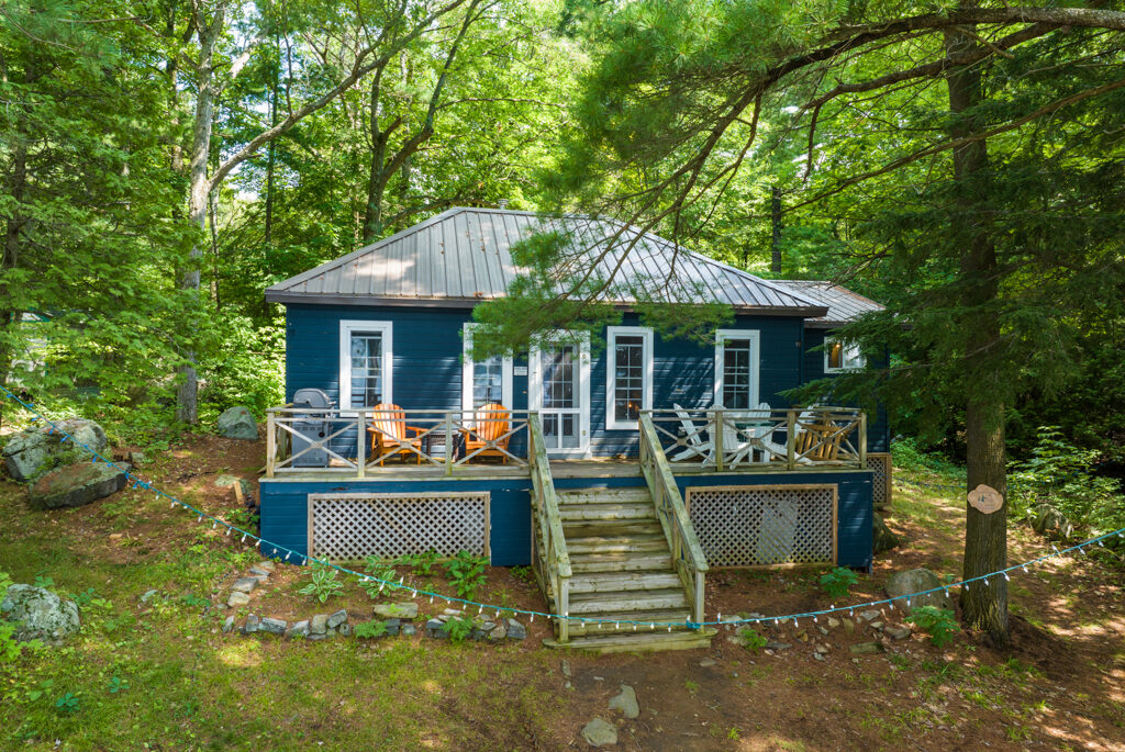 A bungalow cottage with a dark blue exterior sits surrounded by green trees.