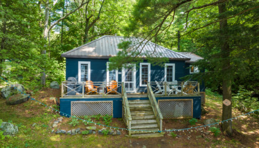 A bungalow cottage with a dark blue exterior sits surrounded by green trees.