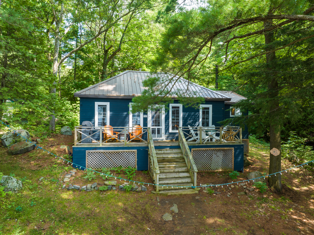 A bungalow cottage with a dark blue exterior sits surrounded by green trees.