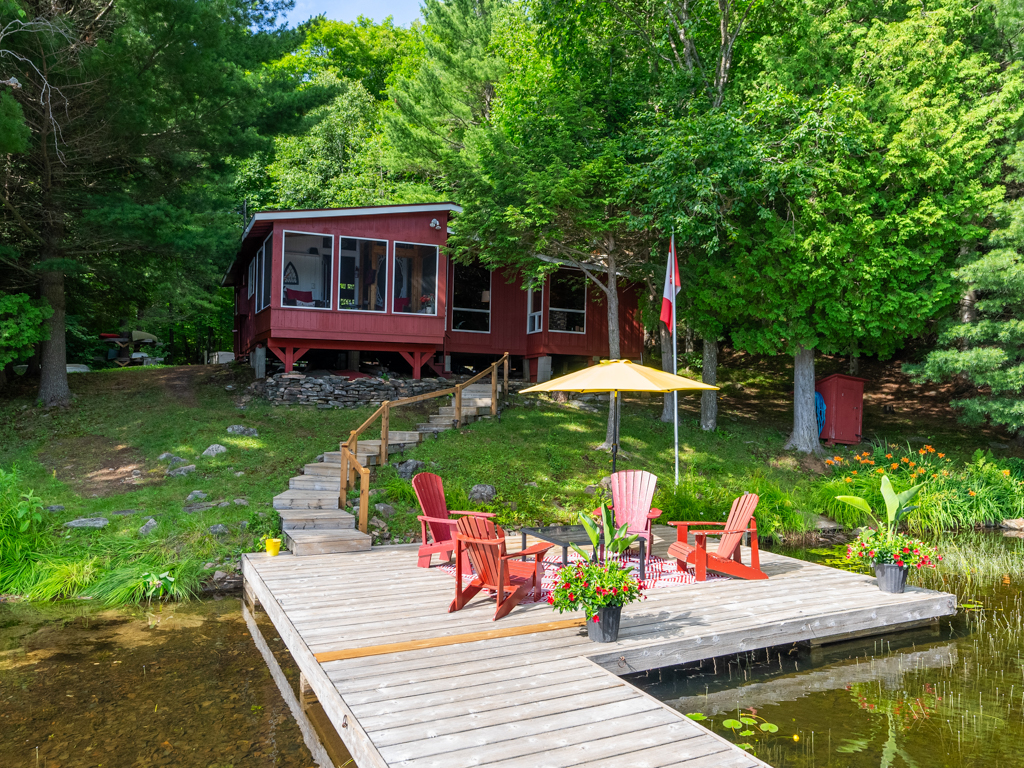 A small red cottage sits just up the shore of a lake, surrounded by green trees. A dock stretches out into the lake in front.