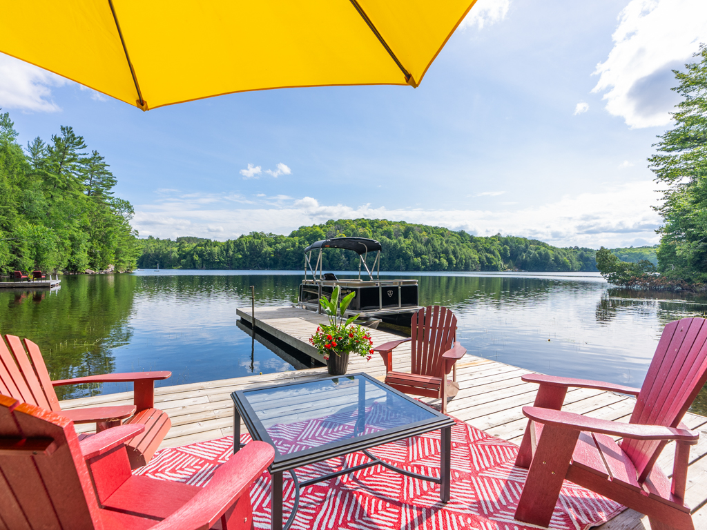 Red patio furniture sits on a wide dock, under a yellow umbrella. A longer piece of dock stretches into the lake, and a pontoon boat sits docked in the water alongside it.
