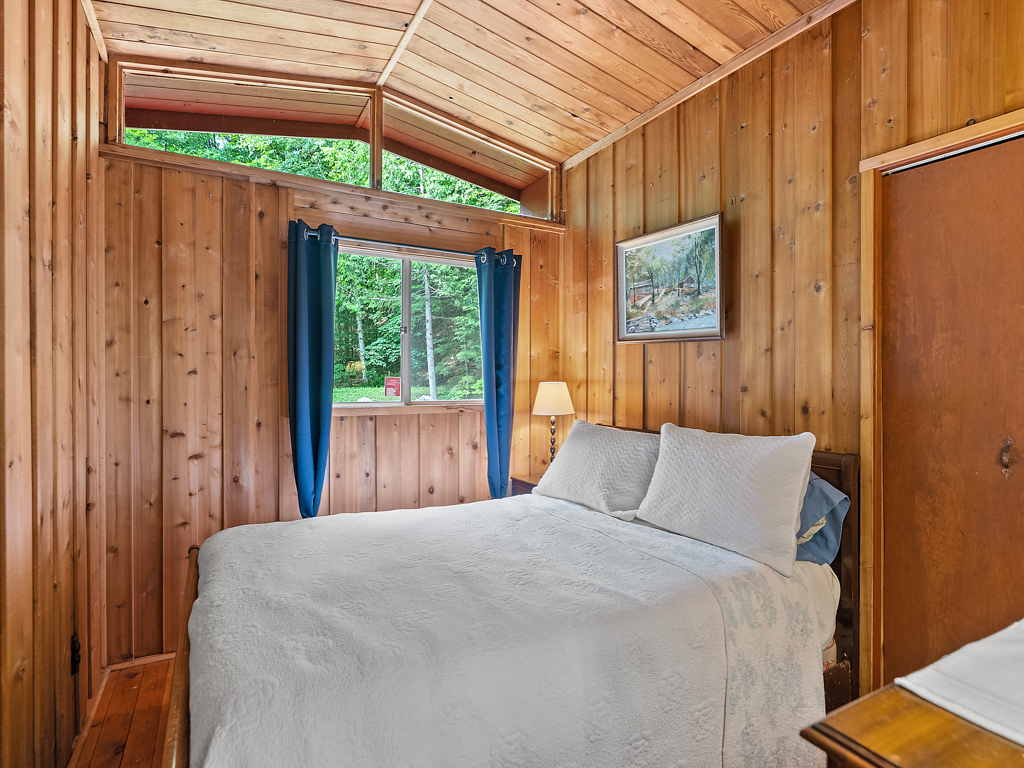 A small bedroom with a white bed, wooden walls and a wooden ceiling, and lots of natural light.