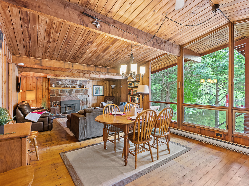 An open-concept cottage interior with a small dining area leading into a living room. Large windows line the lake-facing wall.