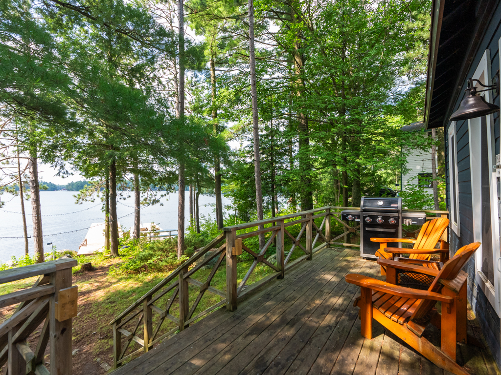 Two wooden Muskoka chairs and a barbecue sit on a deck off the front of a cottage. Stairs lead down off the deck, towards the lake.