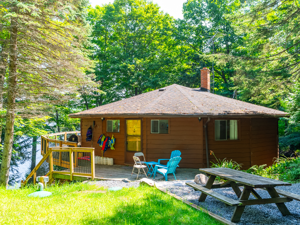 A round cottage with a wraparound deck sits just past a gravel driveway with a picnic table.