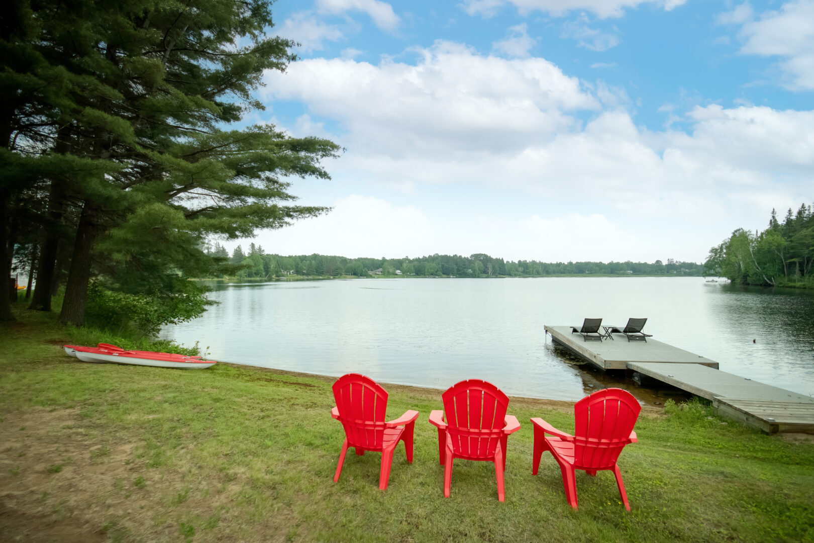 Three red Muskoka chairs sit on a grassy area, facing a still lake.
